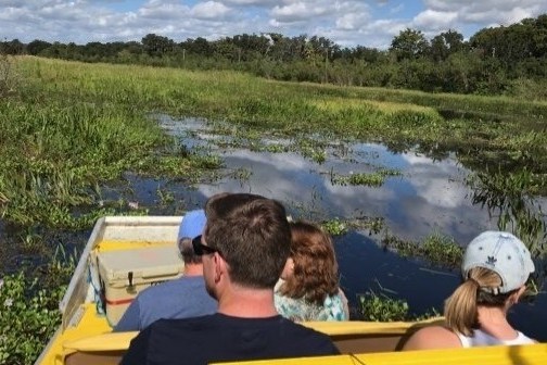 People on an airboat ride through a grassy swamp under a cloudy sky.